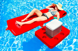 A woman relaxes on a red pool float in a clear blue pool, wearing a hat and bikini, with a cooler beside her. Perfect for summer leisure.