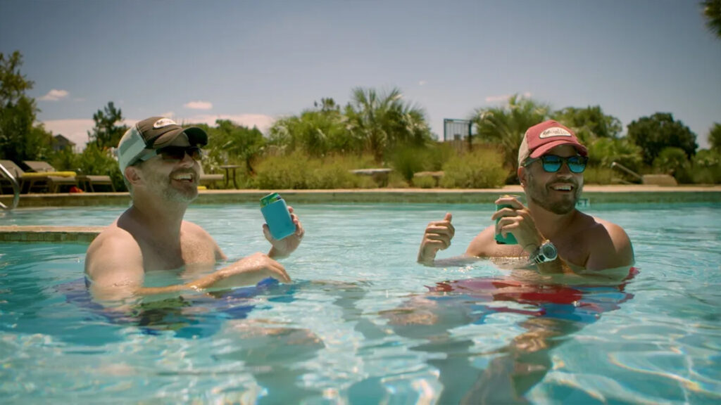 Two men relax in a swimming pool, enjoying drinks and smiling. Lush greenery surrounds the pool, indicating a sunny, leisure-filled day.