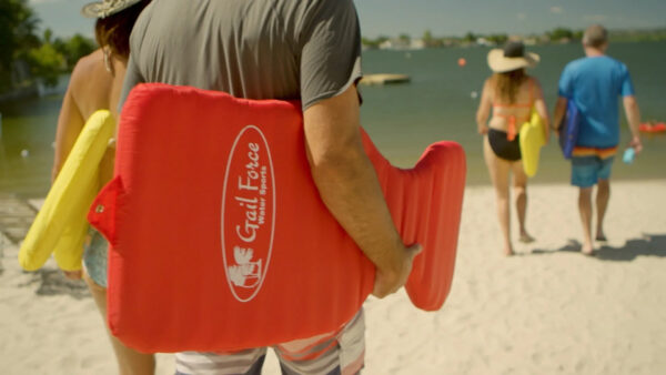 A person carries a red flotation device labeled "Gail Force" towards the water, accompanied by another individual holding a yellow float.